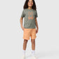 A child with curly hair stands smiling against a light gray background. They wear an olive green KIDS FLOYD GRAPHIC TEE - B0U338B2TS by Psycho Bunny with an orange bunny graphic, peach-colored shorts, white socks, and white sneakers. Their hands rest on their hips.
