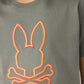 Close-up of a person wearing an olive green KIDS FLOYD GRAPHIC TEE - B0U338B2TS by Psycho Bunny with an orange embroidered bunny outline above two crossed bones on the front. The person appears to have curly hair, partially visible at the top of the image.