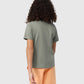 A person with curly hair, seen from the back, is wearing a grey t-shirt with an embroidered Bunny outline and peach shorts. They are standing against a plain, light grey background. The t-shirt they are wearing is the KIDS FLOYD GRAPHIC TEE - B0U338B2TS by Psycho Bunny.
