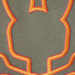 A close-up of fabric with an intricate, abstract design in vibrant orange and red thread on a gray background. The embroidery features bold lines and curves, creating a pattern that appears symmetrical and detailed, resembling an embroidered KIDS FLOYD GRAPHIC TEE - B0U338B2TS by Psycho Bunny outline.
