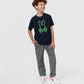 A young boy with curly hair smiles while wearing a dark blue KIDS SANTA MONICA EMBROIDERED GRAPHIC TEE from Psycho Bunny and gray pants, paired with white sneakers. He stands casually against a white background.