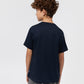 A young person with curly hair wearing a navy blue KIDS SANTA MONICA EMBROIDERED GRAPHIC TEE by Psycho Bunny, viewed from the back, standing against a plain white background.
