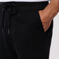A person is wearing black Psycho Bunny MENS OUTLINE SWEATPANT - B6P506ARCN with a drawstring and their left hand in the pocket. There is an embroidered Bunny logo on the pants near the upper thigh, featuring a bunny with crossbones beneath it. The person is also wearing a white shirt.