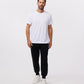 A man with short brown hair and a beard is standing against a plain white background. He is wearing a white T-shirt, MENS OUTLINE SWEATPANT - B6P506ARCN by Psycho Bunny, and white sneakers. His arms are relaxed at his sides, and he is looking straight ahead.