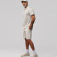 A man with a beard is standing with his hands in his pockets. He is wearing a light gray polo shirt and matching Psycho Bunny MENS OUTLINE SWEATSHORT - B6R507X1CN, a white cap, white socks, and light gray sneakers. The background is plain and light-colored.