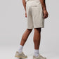 A person stands against a plain background, shown from the waist down. They are wearing MENS OUTLINE SWEATSHORT - B6R507X1CN by Psycho Bunny, ankle socks, and beige sneakers. The person's right arm, partially visible, has a tattoo.