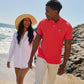 A man in a red Psycho Bunny men's classic polo shirt with the Psycho Bunny logo and a woman in a white shirt and straw hat walk hand in hand along a sunny beach, smiling at each other.