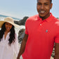 A young man in a red Psycho Bunny Pima cotton polo shirt smiles at the camera on a sunny beach, with a young woman in a white shirt and sun hat walking beside him, waves crashing in the background.