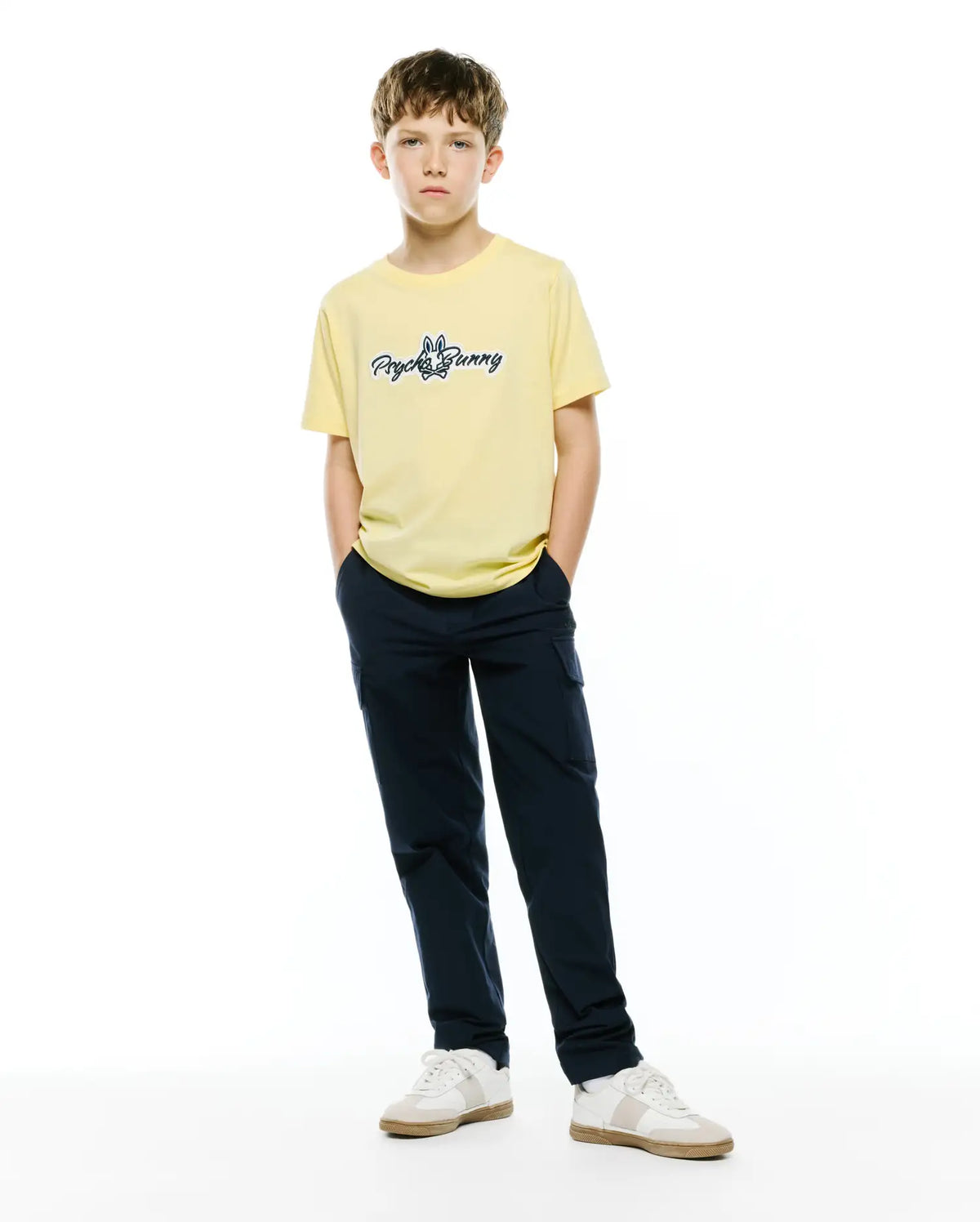 Kids Jaydon Wordmark Graphic Tee, a pale yellow classic fit jersey t-shirt with a medium blue embroidered Psycho Bunny wordmark logo on the center chest, styled with navy cargo pants. (4)
