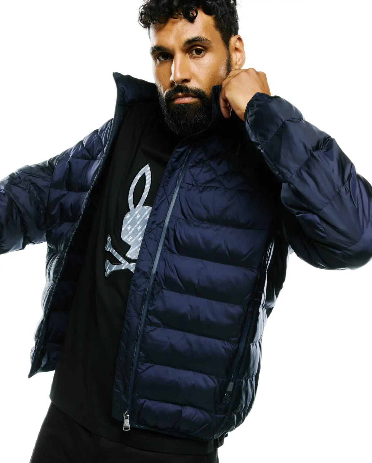 Mens Emerik Hooded Puffer Jacket in dark navy, with horizontal quilting and full zip. Layered over a black jersey t-shirt featuring a large grey Psycho Bunny skull logo with subtle pattern on the chest. (3)