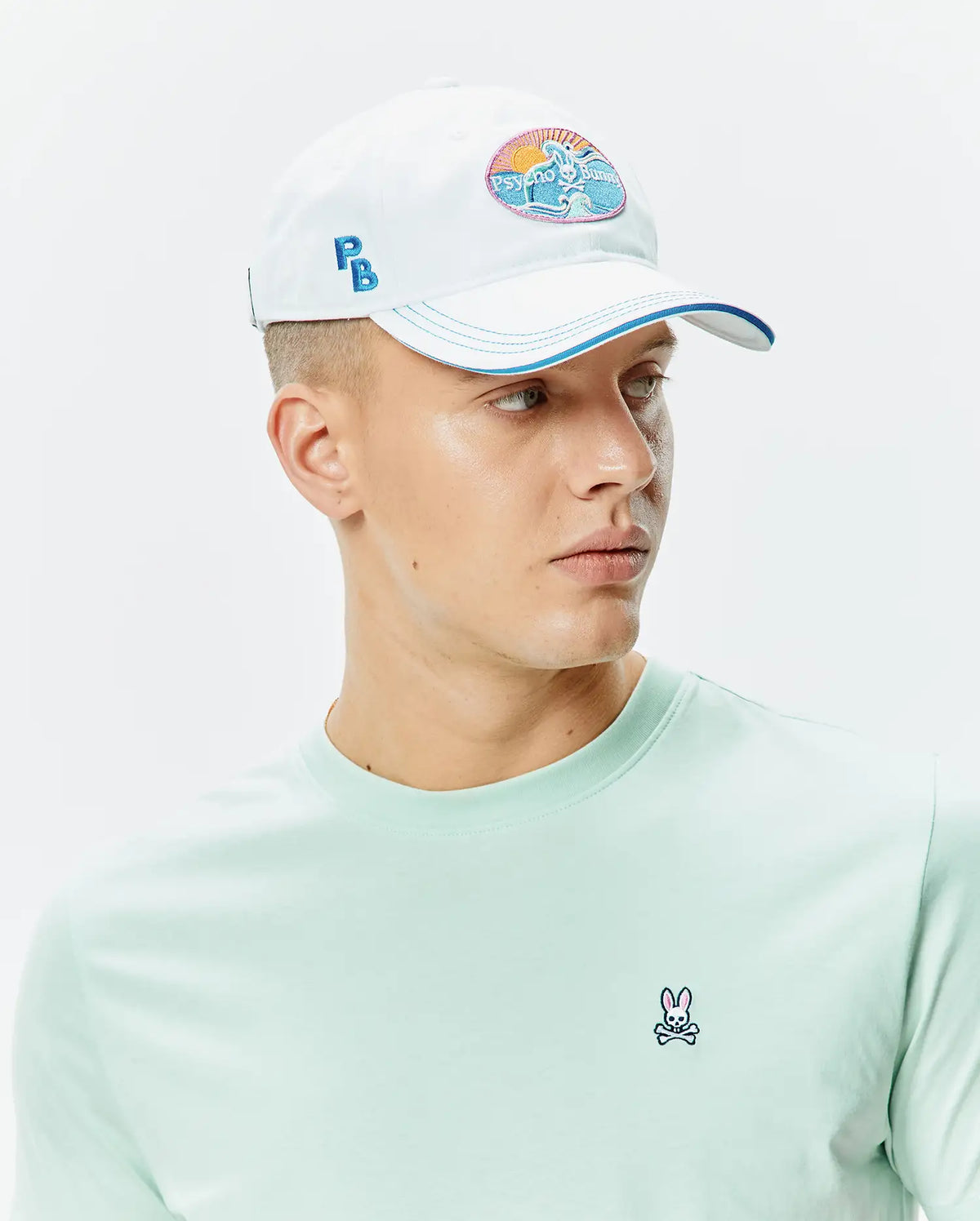 White Mens Ray Baseball Cap with a Psycho Bunny beach patch, blue PB embroidery, and blue brim, alongside a light green jersey crew neck t-shirt with a small embroidered Psycho Bunny skull on the left chest. (1)