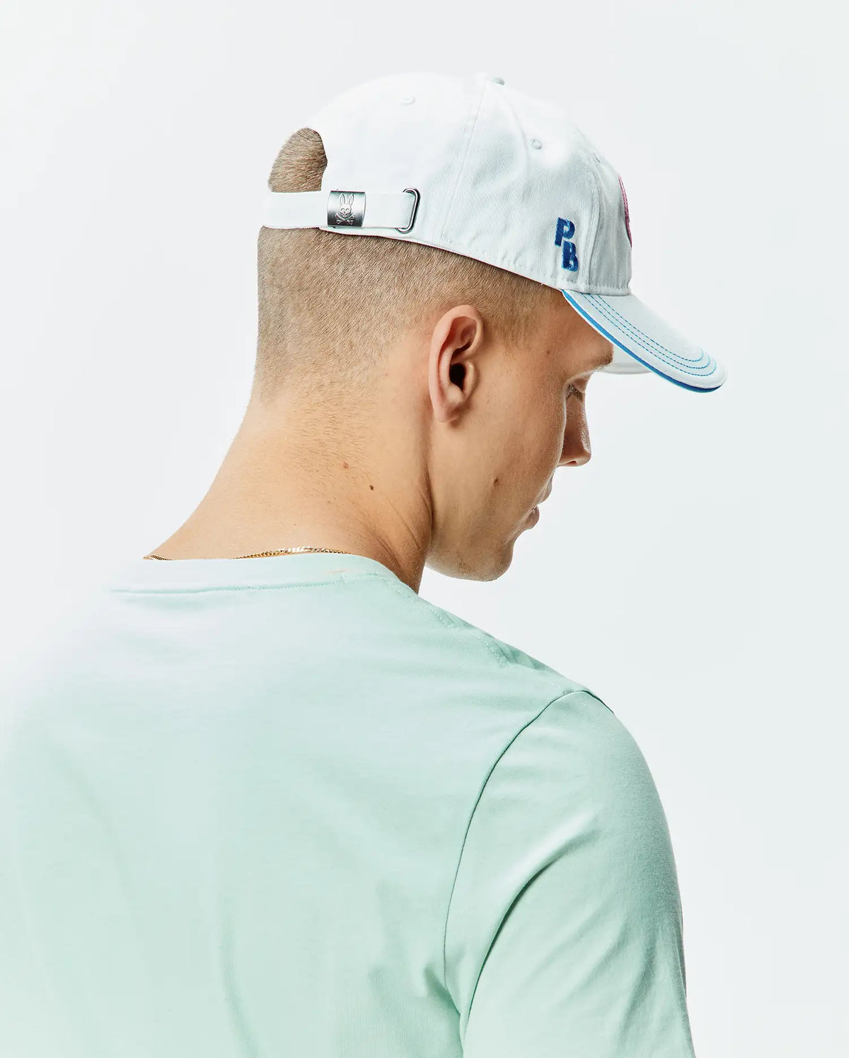Pale blue Mens Ray Baseball Cap with blue brim stitching, embroidered blue PB initials, a pink Psycho Bunny skull logo, and a metal buckle with engraved bunny skull, paired with a light mint green jersey t-shirt. (3)