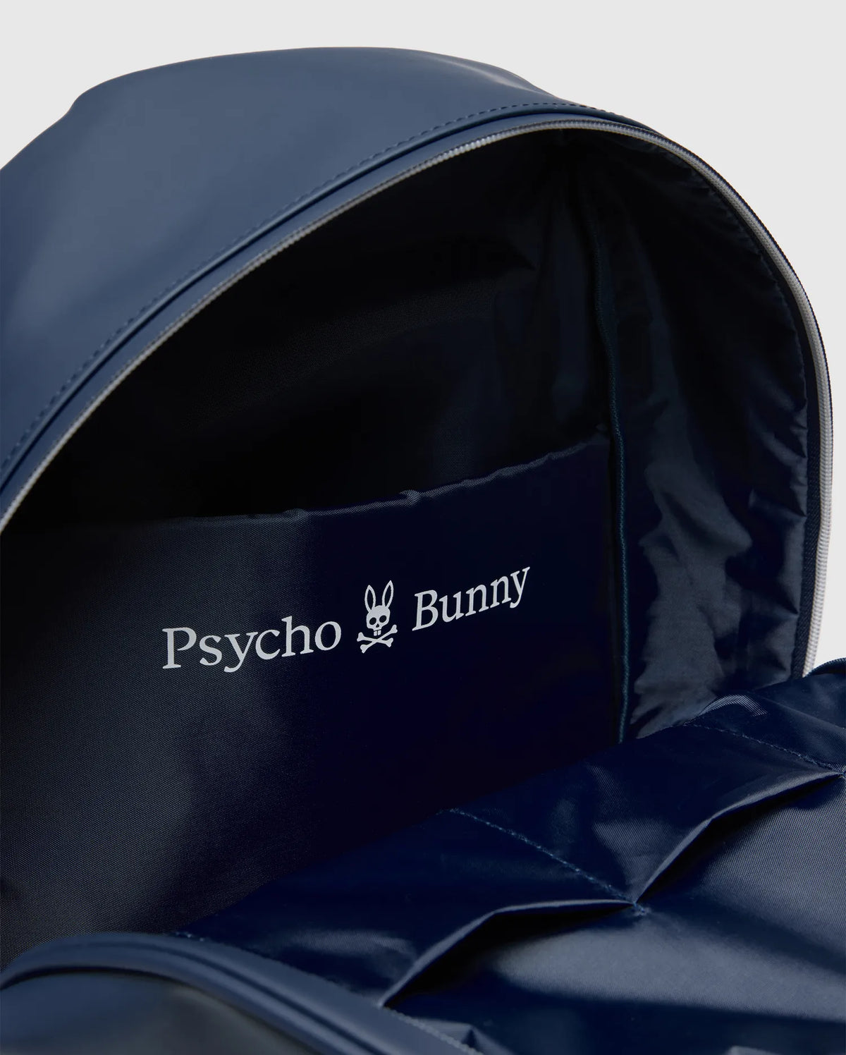Classic Bunny Backpack in navy blue, featuring a smooth exterior, light grey zipper, and a prominent white Psycho Bunny skull logo and text print on an interior lining pocket with multiple slip pockets. (3)