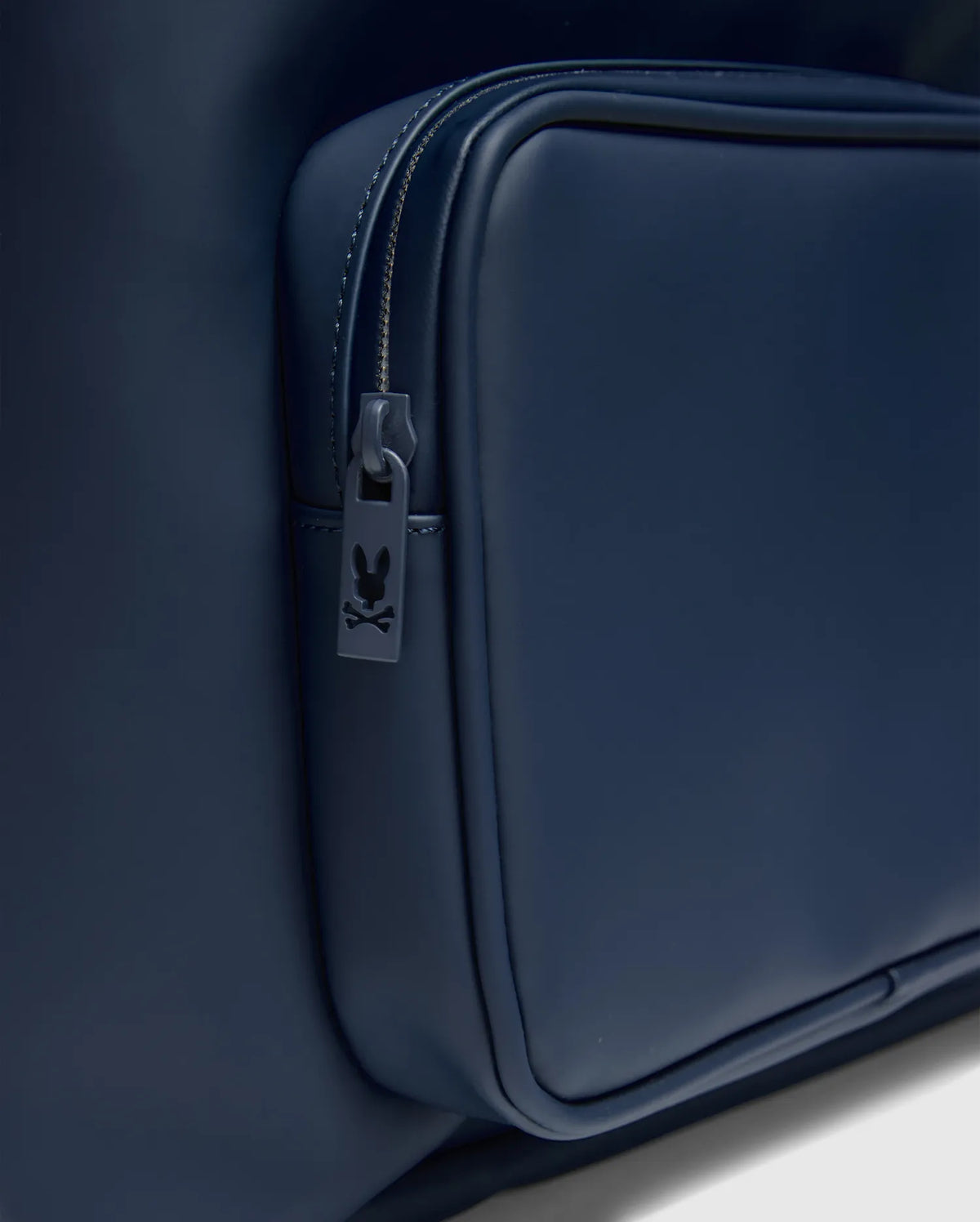 Classic Bunny Backpack in navy blue with a smooth, matte finish, featuring a zippered front pocket and a small Psycho Bunny skull logo on the zipper pull. (4)