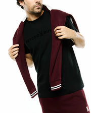 Mens Royce Graphic Tee, a black jersey t-shirt with a subtle black Psycho Bunny text and skull logo chest graphic, styled with burgundy shorts featuring a white embroidered bunny and a draped striped-cuff sweatshirt. (4)