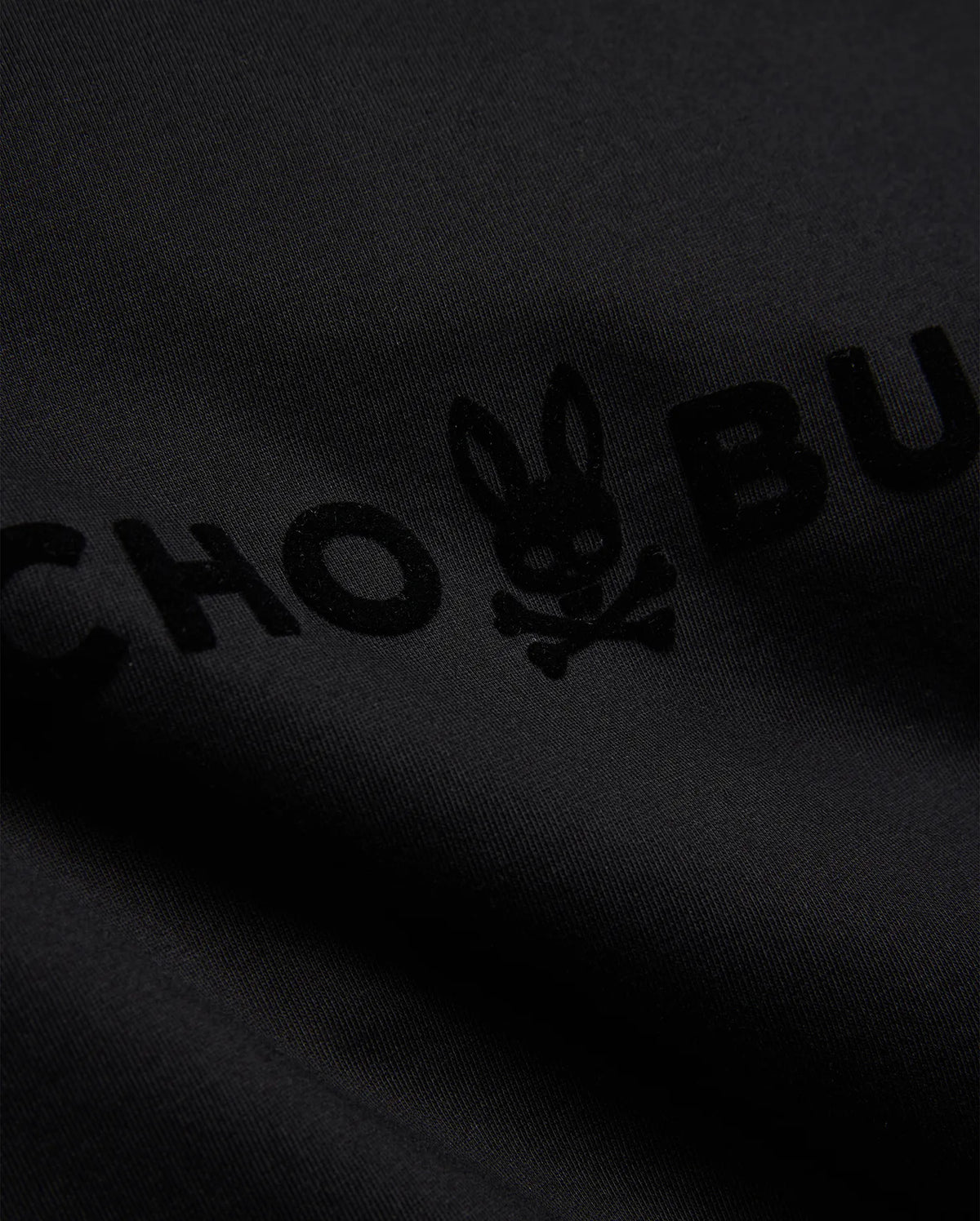 Mens Royce Graphic Tee, a black jersey knit t-shirt featuring a prominent, tonal black velvet-textured Psycho Bunny text and iconic bunny skull logo graphic. (6)