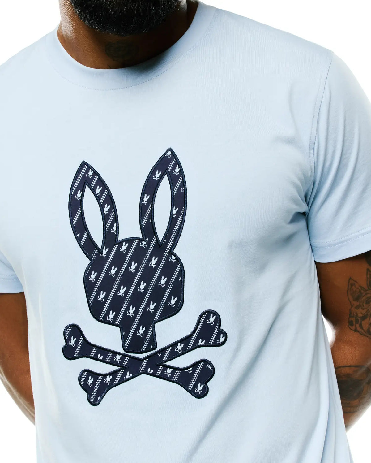 Light blue Mens Jaden Appliqué Graphic Tee, classic fit, smooth jersey fabric. Features a large navy and white patterned Psycho Bunny skull logo appliqué on the chest with small bunny skulls and crossbones. (3)