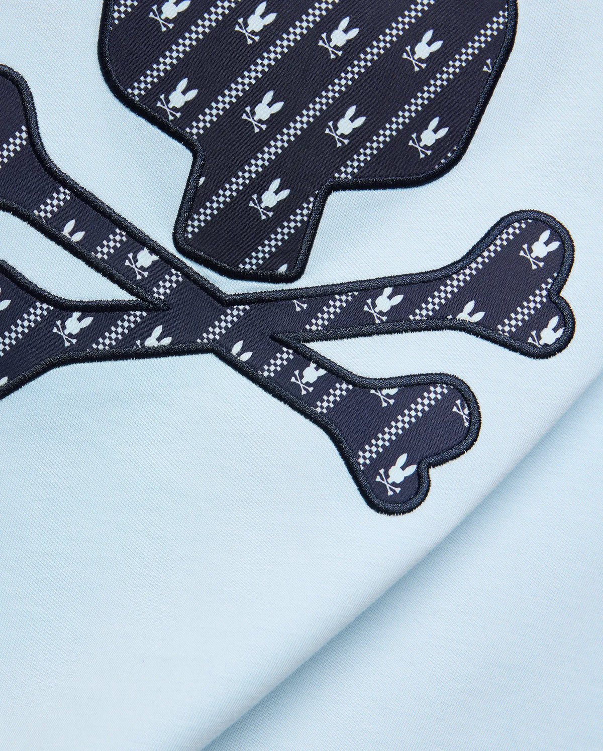 Mens Jaden Graphic Tee in light blue jersey knit, featuring a large navy blue skull and crossbones graphic with white Psycho Bunny logo and checkered stripe pattern, outlined in black embroidery. (6)