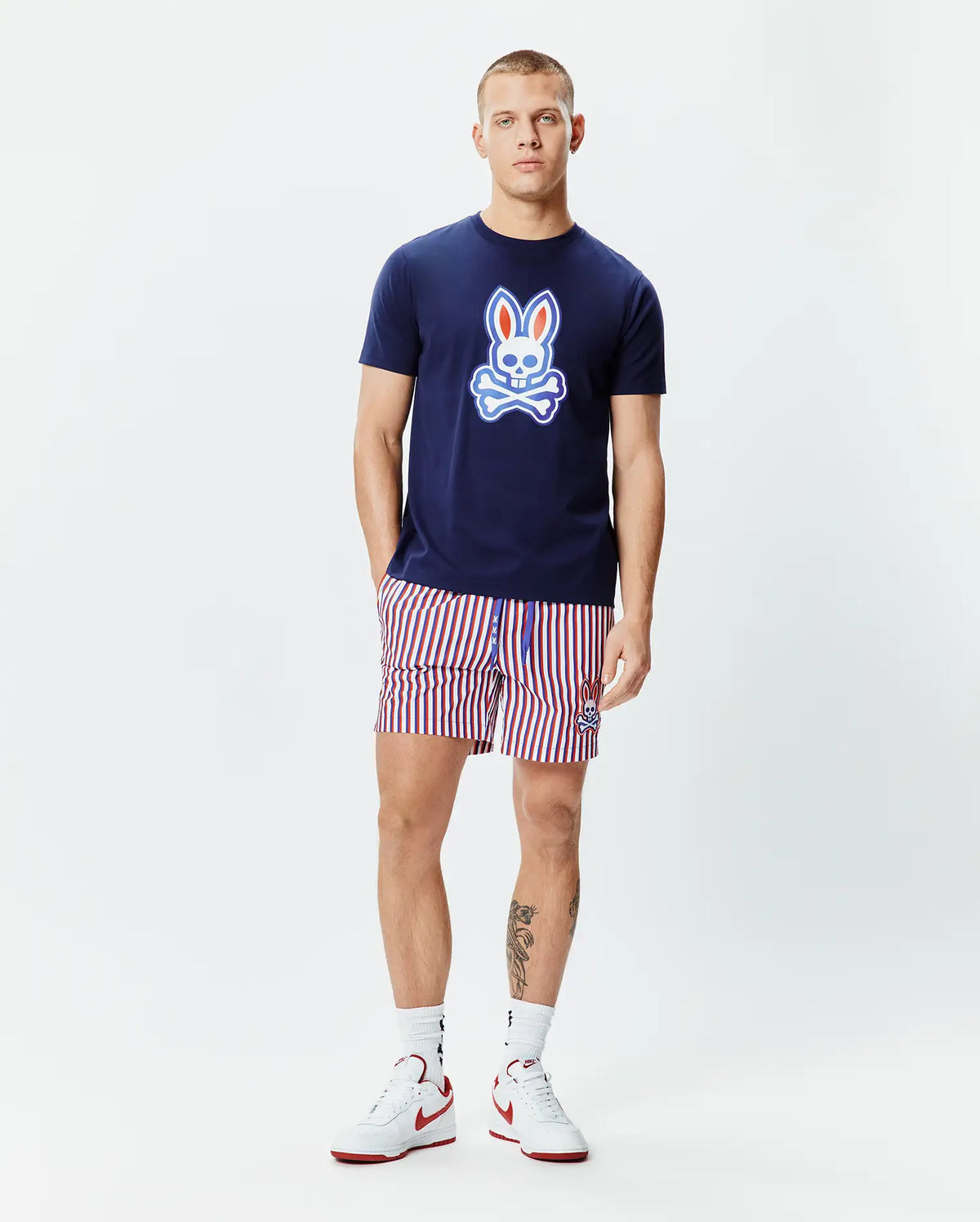 Mens John Graphic Tee, a navy blue jersey t-shirt with a large red, white, and light blue Psycho Bunny skull logo on the chest, paired with red, white, and navy striped shorts featuring a small embroidered bunny logo. (5)