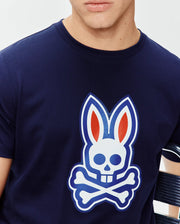 Mens John Graphic Tee, a navy blue classic fit jersey knit t-shirt featuring a large central chest graphic of the iconic Psycho Bunny skull logo in white, blue, and red with an embroidered outline. (4)