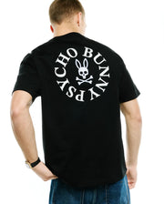 Mens Olek Relaxed Back Graphic Tee, black jersey knit, featuring a large white circular Psycho Bunny skull and crossbones logo with text on the back. (1)