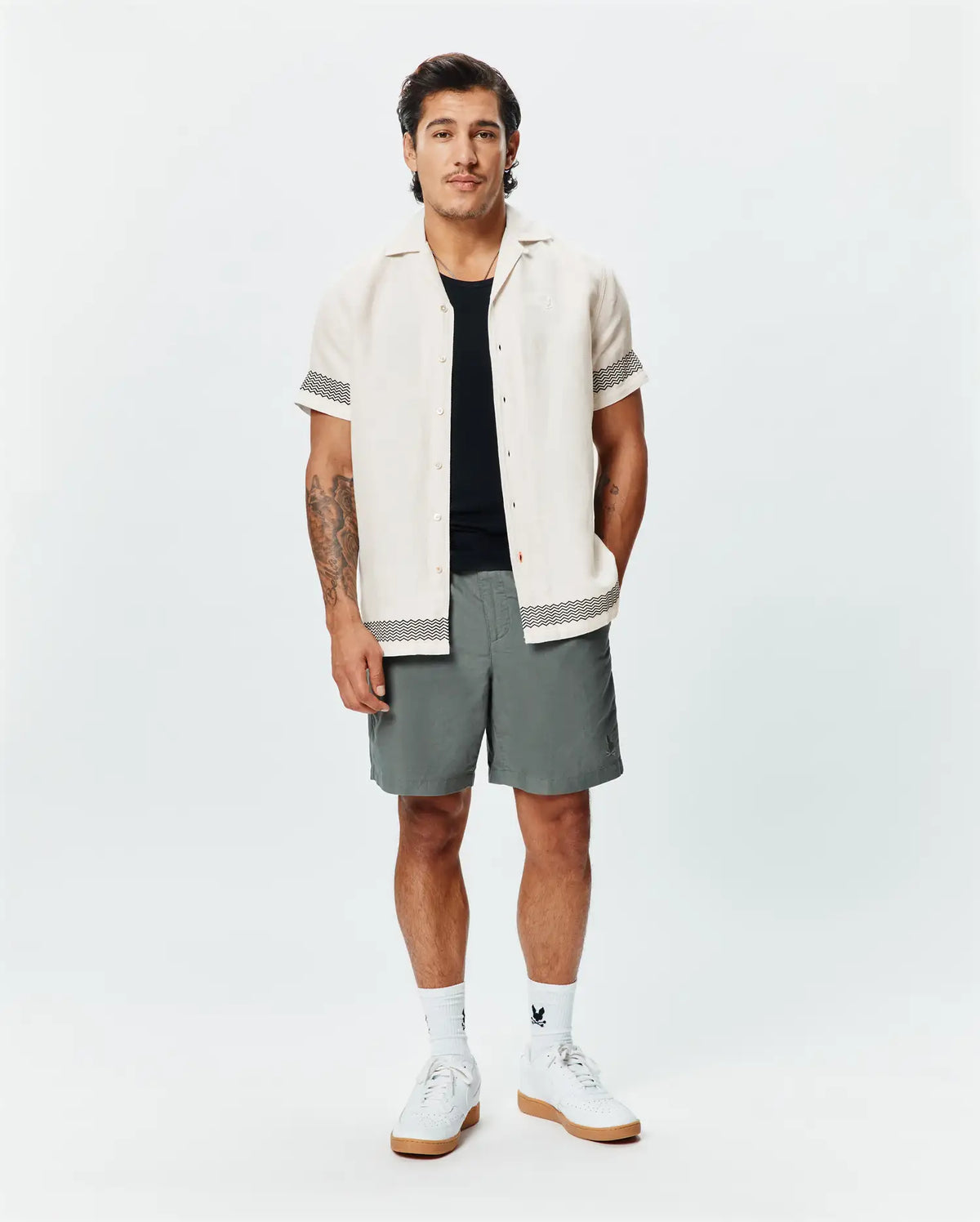 Mens Sunny Linen Shirt, cream, with black zigzag trim on sleeves and hem, paired with grey shorts featuring a small Psycho Bunny logo, white crew socks, and white sneakers. (4)