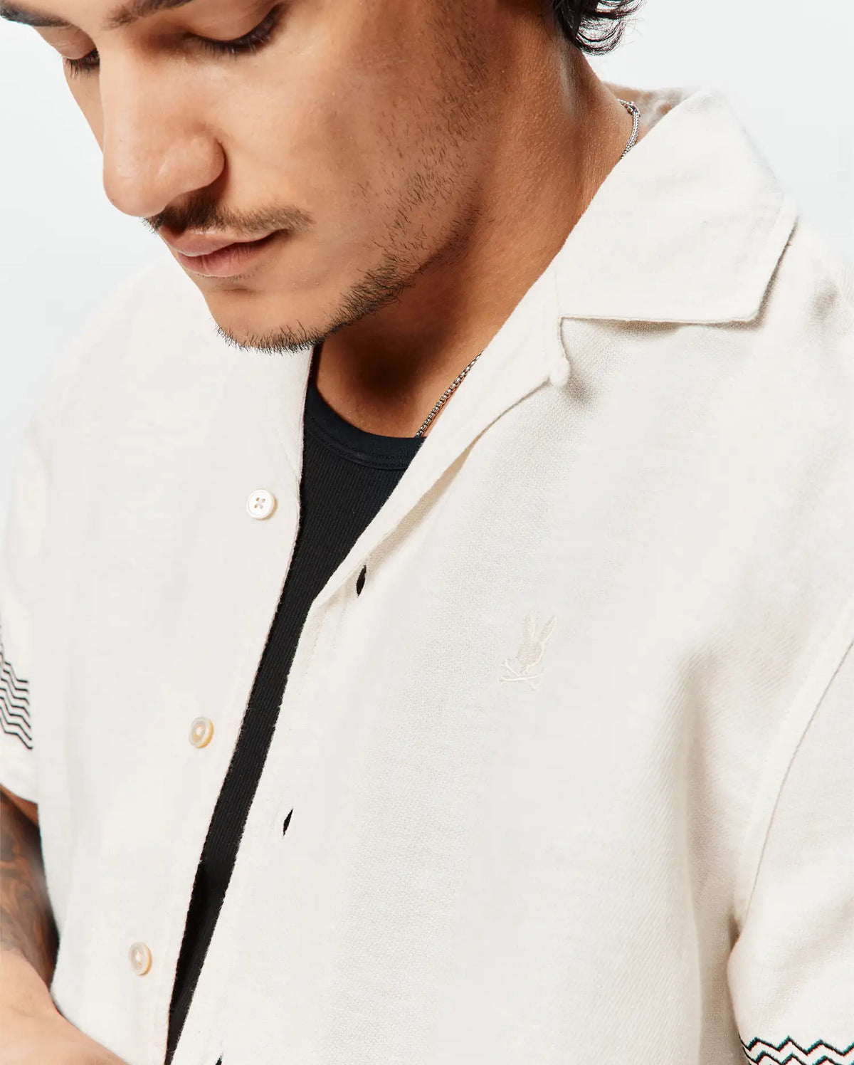 Mens Sunny Linen Shirt, a relaxed fit off-white linen camp collar shirt featuring a small embroidered Psycho Bunny skull logo on the left chest and black and teal chevron sleeve pattern. (2)