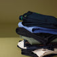 A neatly stacked pile of various pieces of clothing, including Psycho Bunny men's Gable regular fit sport pants and shirts, on a green background. The clothes feature subtle, stylish stitching and detailing.