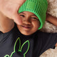 A young child wearing a bright green beanie and a black Psycho Bunny Pima cotton t-shirt with an embroidered green bunny logo, smiling and lying on a fluffy white surface, partially covering their face with a hand.