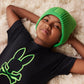 A young child lies on a fluffy white rug, wearing a black Psycho Bunny Pima cotton t-shirt featuring a neon green bunny embroidered design. They are smiling and wearing a bright green knitted hat.