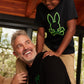 A joyful scene featuring an older man with gray hair and a younger person in a green beanie, both smiling and wearing black Psycho Bunny graphic tees with a neon green bunny logo, in a cozy wooden cabin setting.