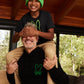 A joyful child in a green beanie and Psycho Bunny KIDS SANTA MONICA EMBROIDERED GRAPHIC TEE covers the eyes of a smiling man with a white beard, who is carrying the child on his shoulders inside a cozy, wooden cabin.