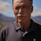An older man with a mustache and short gray hair, wearing a dark Psycho Bunny Men's Classic Polo - B6K001ARPC shirt with a small logo, stares intensely at the camera against a blurred landscape background.