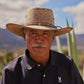 An older man wearing a straw hat and a dark, crease-resistant Psycho Bunny collar polo stands outdoors, with a calm expression and desert vegetation in the background.