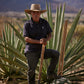 An older man wearing a Psycho Bunny Pima cotton polo and straw hat stands with a wooden stick next to an agave plant in a sunny, mountainous landscape.