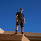 A man wearing black Psycho Bunny OUTLINE sweat shorts and sunglasses stands confidently at the top of sandy concrete steps under a clear blue sky.
