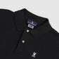 Close-up image of a black Psycho Bunny Pima cotton polo shirt with a buttoned, crease-resistant collar, and an embroidered white bunny logo on the left chest area. The inner collar label is visible showing MENS CLASSIC POLO - B6K001ARPC.