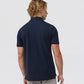 A person with wavy blonde hair is shown from behind, wearing a navy blue Psycho Bunny MENS CLASSIC POLO - B6K001ARPC made of diamond-knit piqué fabric and light gray pants. The background is plain white. A small tattoo is visible on the person's left forearm.