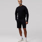 A man with gray hair and beard stands confidently, dressed casually in a black MENS OUTLINE SWEATSHIRT made from premium textiles, black shorts, white socks, and white sneakers against a neutral gray background by Psycho Bunny.