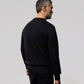 A man with grey hair stands facing away from the camera, wearing a plain black Psycho Bunny MENS OUTLINE SWEATSHIRT - B6S503ARCN and black pants against a light grey background.