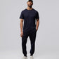 A man stands against a plain background, wearing a light blue cap, dark jogger pants, and white sneakers. He has a neutral expression and his hands are relaxed by his sides. He is wearing a navy blue Psycho Bunny MENS OUTLINE TEE - B6U500ARPC made from Pima cotton.