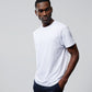 A man stands against a plain background, wearing a white MENS OUTLINE TEE - B6U500ARPC from Psycho Bunny and dark pants. His right hand is in his pocket while his left arm hangs by his side. He looks slightly to his right with a relaxed expression.