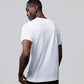 A man with short hair is standing and looking over his shoulder. He is wearing a plain white MENS OUTLINE TEE - B6U500ARPC from Psycho Bunny and dark pants from the OUTLINE collection. The background is a light gray color.