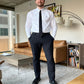 A man in a white shirt and Psycho Bunny men's Gable sport pants stands confidently in a modern living room, wearing a black tie and dress shoes.