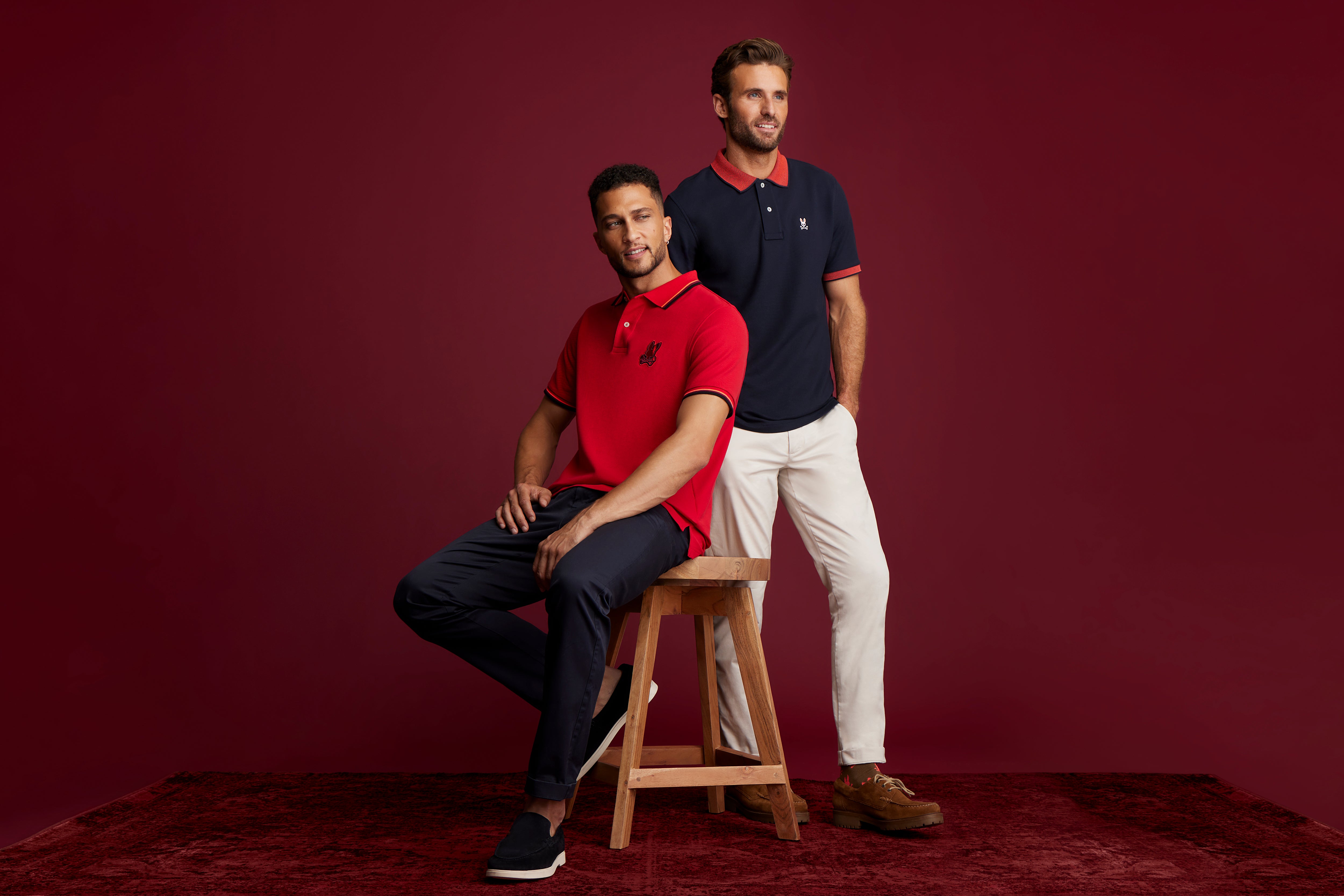 Two men pose against a burgundy backdrop; one sits on a wooden stool wearing a red polo and dark pants, while the other stands in a navy polo and white pants. Both are smiling.
