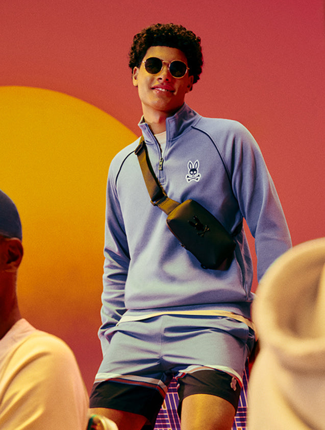 A young man in a red plaid shirt leans out of the window of a vintage green car, smiling as he reaches down to pick up a football from the ground.