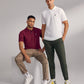 Two men posing in a studio, one sitting on a cube wearing a burgundy Psycho Bunny MENS CLASSIC POLO - B6K001ARPC and beige pants, the other standing in a white polo and green pants. Both are wearing white sneakers.