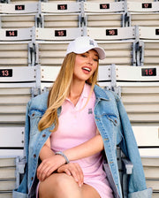A woman with long blonde hair wearing the Psycho Bunny WOMENS POLO DRESS - WO2000003 and a denim jacket sits smiling in stadium seats 14 and 15, her feminine silhouette highlighted as she faces the camera.