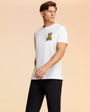 A man with short brown hair stands against a beige background, wearing a Psycho Bunny MENS VEGAS CITY TEE - B6U967X1PC made of soft Pima cotton jersey. The white t-shirt features a yellow and black printed bunny logo on the chest. He is also wearing black pants and looking to his left with a neutral expression.
