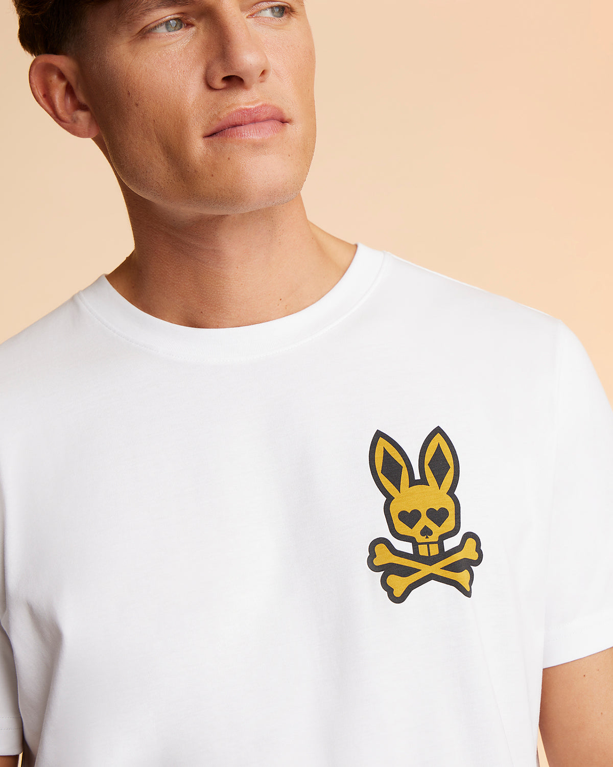 A person wearing a white Psycho Bunny MENS VEGAS CITY TEE - B6U967X1PC with a distinctive printed Bunny Logo featuring a cartoonish rabbit skull and crossbones on the upper left side is shown. The background is a gradient of warm, peachy tones. The person has short hair and is looking off to the side.